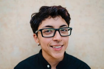 close up view of a young transgender man with black hair and glasses looking at the camera and smiling